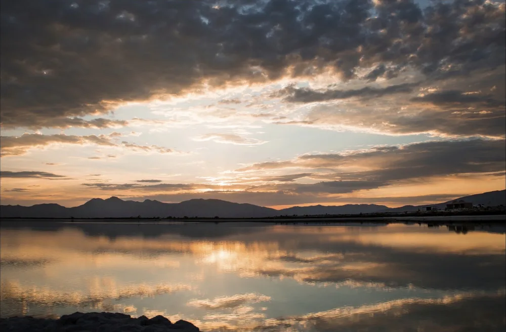 Untitled photograph by Mariana Yáñez — a still mountain lake at dusk mirroring a cloud-filled sky lit gold and blue by the setting sun.