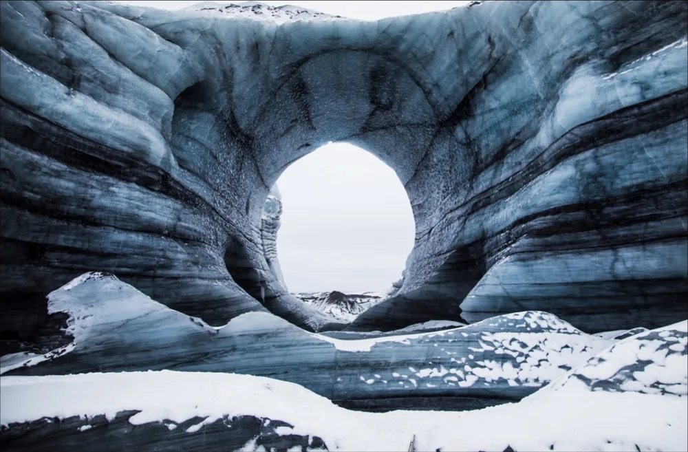 Untitled photograph by Mariana Yáñez — the sculpted interior of a blue glacial ice cave opening to daylight through a rounded portal above snow.